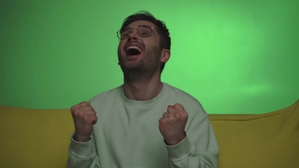 Aufgeregter Mann Mit Brille Zeigt Siegergeste Beim Fernsehen Auf Grün — Stockvideo