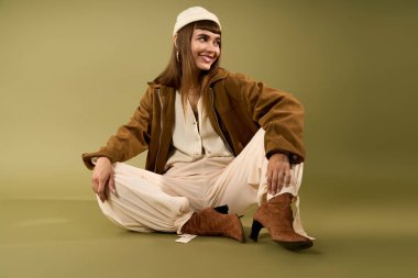 Moda kıyafetleri giymiş neşeli genç bir kadın yeşil bir arka planda gelişigüzel oturuyor ve neşe saçıyor..