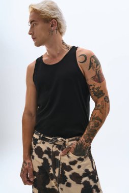 Dövmeleri olan genç bir adam, sade bir arka planda, modaya uygun bir kıyafetin içinde duruyor..