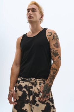 Modayı takip eden genç adam kendine güvenerek poz veriyor. Parlak bir zemine karşı kıyafetlerini ve dövmelerini sergiliyor..