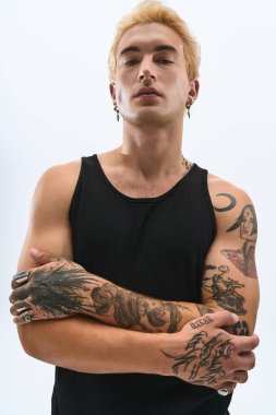 Yakışıklı genç bir adam kollarını kavuşturup şık kıyafetlerini ve karmaşık dövmelerini gösteriyor..