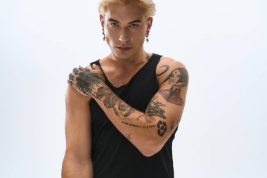 Genç adam kollarını kavuşturmuş, dövmelerini ve şık kıyafetlerini sergiliyor..