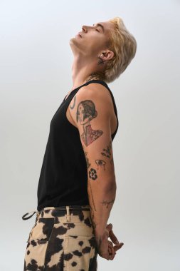 Genç adam kendine güvenerek poz veriyor, dövmelerini ve moda tarzını bir sanat stüdyosunda vurguluyor..