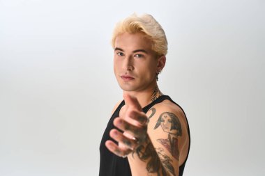 Dövmeli şık genç adam elini uzatıyor, minimalist bir yerde poz veriyor..