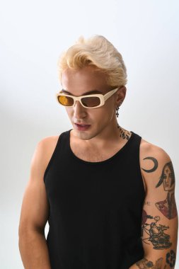 Delikanlı, gösterişli dövmelerini ve şık gözlükleriyle gösteriş yapıyor. Düz bir zemine karşı şık bir kıyafet giyiyor..