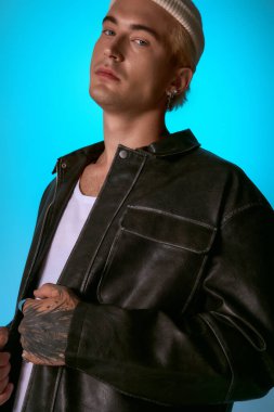 Yakışıklı genç adam şık bir ceketle poz veriyor ve parlak mavi bir arkaplan karşısında havalı bir tavır sergiliyor..