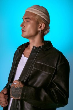 Zarif genç adam, parlak mavi bir dekor içinde parlak bir ceket ve şapka giymiş, yetenekli poz verir..