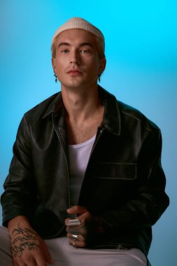 Yakışıklı genç adam kendine güvenerek poz veriyor, parlak bir deri ceket ve şık aksesuarlar giyiyor..