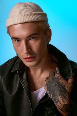 Kendine güvenen genç bir adam, kendine has bir tarzla poz verir. Moda kıyafetleri ve çarpıcı dövmeleri vardır..