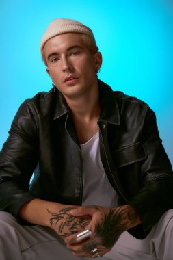 Dövmeleri olan genç bir adam kendine güvenerek şık bir şekilde poz veriyor ve kendine has tarzını ön plana çıkarıyor..