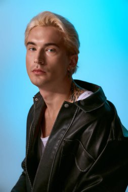 Yakışıklı genç adam siyah deri ceketle havalı mavi bir arka planda rahatça poz veriyor..