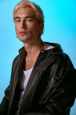 Yakışıklı genç bir adam, açık mavi arka planı olan modaya uygun siyah deri bir ceketle güven içinde poz veriyor..