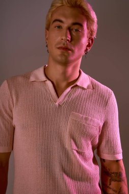 Sarı saçlı genç bir adam, parlak ışıkla aydınlatılan modaya uygun giyinmiş, kendinden emin bir şekilde poz veriyor..