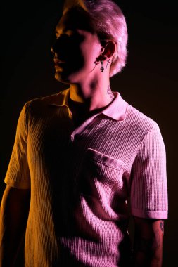 Yakışıklı genç adam kendine güvenerek poz veriyor, dramatik ışıklandırma altında moda anlayışını vurguluyor..