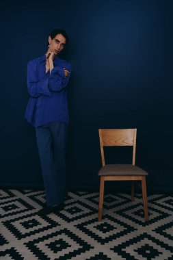 Şık bir adam ahşap bir sandalyenin yanında duruyor, keskin stilini ve zarafetini sergiliyor..