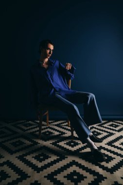 Modaya uygun mavi elbiseler giyen yakışıklı adam loş bir odada rahatça poz veriyor..