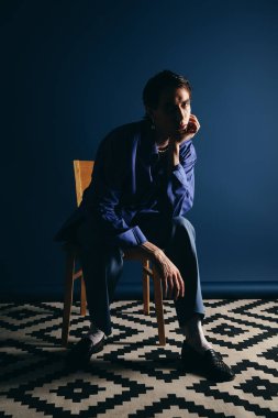 Yakışıklı bir adam düşünceli bir şekilde bir sandalyeye oturmuş, sakin bir ortamda eşsiz bir moda sergiliyor..