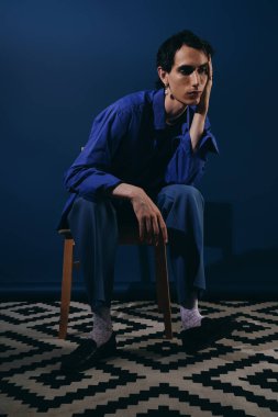 Yakışıklı bir adam ahşap bir sandalyede poz veriyor, mavi bir arka plana karşı moda anlayışını sergiliyor..