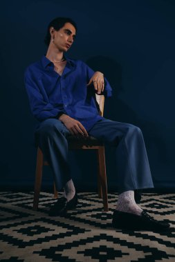 Kobalt mavisi gömlekli yakışıklı bir adam zarif bir şekilde bir sandalyede oturur..
