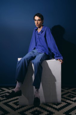 Yakışıklı bir adam kürsüde otururken modern bir stil sunar, şık kıyafetini vurgular..