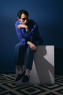 Şık giyinmiş yakışıklı bir adam mavi bir duvara karşı derin düşüncelere dalmış bir kaidede oturuyor..