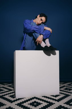 Mavili şık bir adam beyaz bir kaidede zarif bir şekilde oturur..