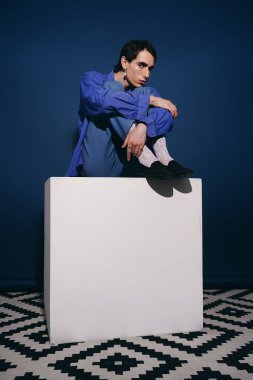 Modaya uygun bir adam beyaz bir platformda oturuyor, şık kıyafetlerini ve çarpıcı duruşunu sergiliyor..