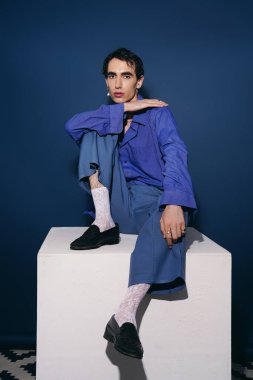 Mavi takım elbiseli yakışıklı bir adam zarif bir kaidede oturuyor çarpıcı bir zemine karşı..