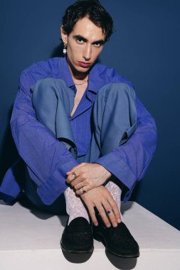 Yakışıklı bir adam zarif bir şekilde katlanmış bacaklarıyla oturur, serin mavi bir elbise giyer ve ışık saçar..