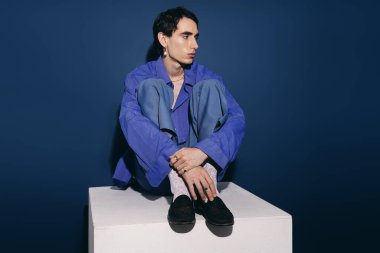 Şık genç adam kendine güvenen bir bakışla bir kaidede oturuyor, modern moda seçimleri sergiliyor..