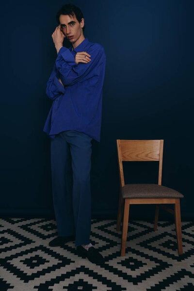Стильный мужчина в ярко-синем наряде стоит рядом со стулом..