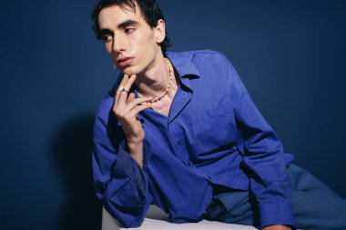 Zarif bir adam yumuşak bir zemine yaslanır, mavi elbisesi zarif bir şekilde arka planla çelişir..