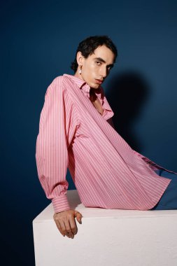 Büyük çizgili gömlekli yakışıklı adam canlı mavi bir duvara karşı kendine güvenen bir duruş sergiliyor.