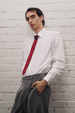 Şık giyinmiş kendine güvenen bir adam beyaz tuğladan bir duvarın yanında duruyor ve kendine has moda anlayışını sergiliyor..