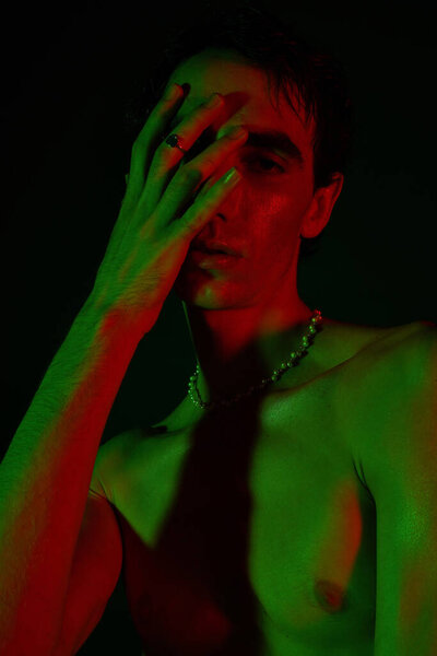 A handsome man poses confidently, partially obscured by his hand, surrounded by striking colors.