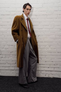 Parlak giyinmiş, uzun paltolu yakışıklı bir adam minimalist bir duvarın karşısında duruyor..