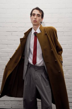 Yakışıklı bir adam uzun paltolu şık bir elbise giyer. Tuğladan bir duvara yaslanır..