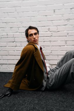 Tarz sahibi bir adam beyaz bir duvar ve siyah bir halıya karşı gelişigüzel poz verir..