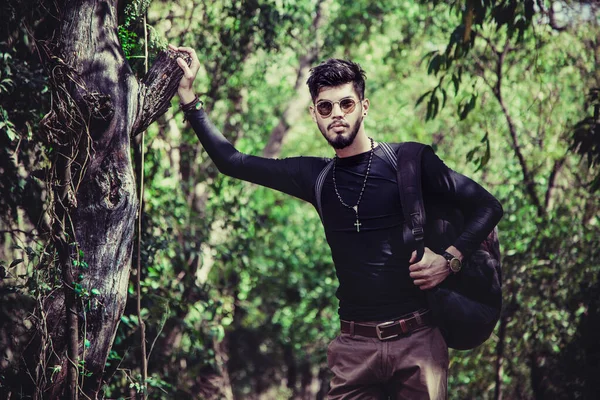 Bir eliyle ağacı tutan adam kameraya bakıyor. Boynunda çapraz zincir, clack tişörtü, kahverengi kemer ve pantolon, saat ve bilezik güneş gözlüğü. Ormanda daha yeşil