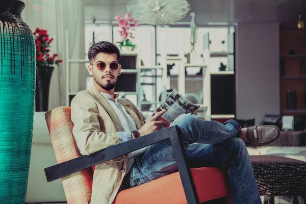 Süslü bir yerde oturan genç ve şık bir adam. İç mekan portresi. Kot pantolon ve bej ceket giyen bir manken. şarjörü elinde tut