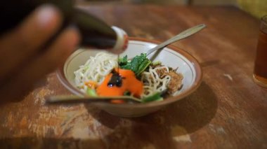 Bir kase Endonezya tavuğuyla sebzeleri karıştıran bir adam. Tahta bir masada çatal ve kaşıkla.