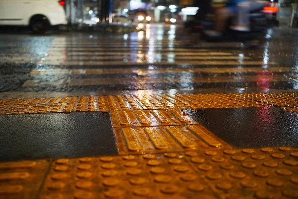 Yağmurlu bir gecede, şehir ışıklarının yansımaları ve ıslak kaldırımları ile çarpıcı bir kentsel atmosfer oluşturan yaya geçidinin yakın görüntüsü.