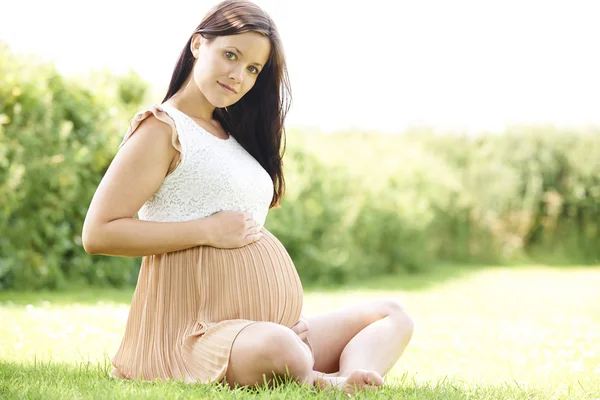 Těhotná žena sedí na trávě, venku drží bump — Stock fotografie