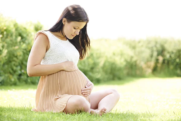 Těhotná žena sedí na trávě, venku drží bump — Stock fotografie