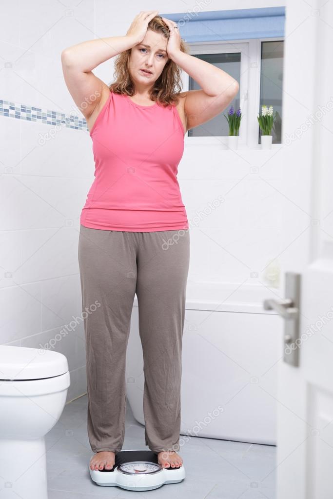 Unhappy Overweight Woman Weighing Herself On Scales In Bathroom