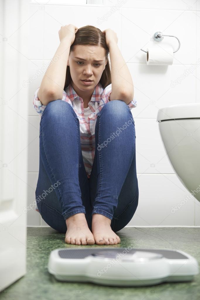 Unhappy Teenage Girl Sitting On Floor Looking At Bathroom Scales