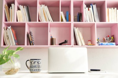Beyaz ofis masasında dizüstü bilgisayarla sabahları bir fincan kahve ve arkasında pembe bir kitaplık.