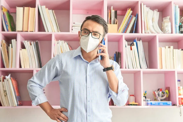 half-length portrait of young latin man with mask holding a smartphone, talking on the phone, young businessman with glasses using the phone in an office in the morning