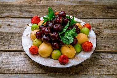 Beyaz tabakta meyve, kiraz, kayısı, çilek, eski ahşap masada nane.