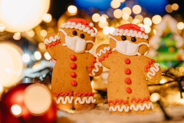 Coronavirus salgını, odaklanmamış ışıklarla kavramsal arka plan Noel süsleri ve koruyucu bir tıbbi maske takan Noel Baba şeklinde zencefilli ekmek.